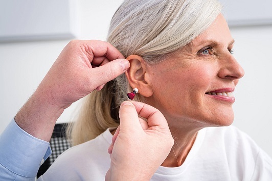 Eine ältere Frau bekommt ein modernes Hörgerät ins Ohr eingesetzt und lächelt dabei.